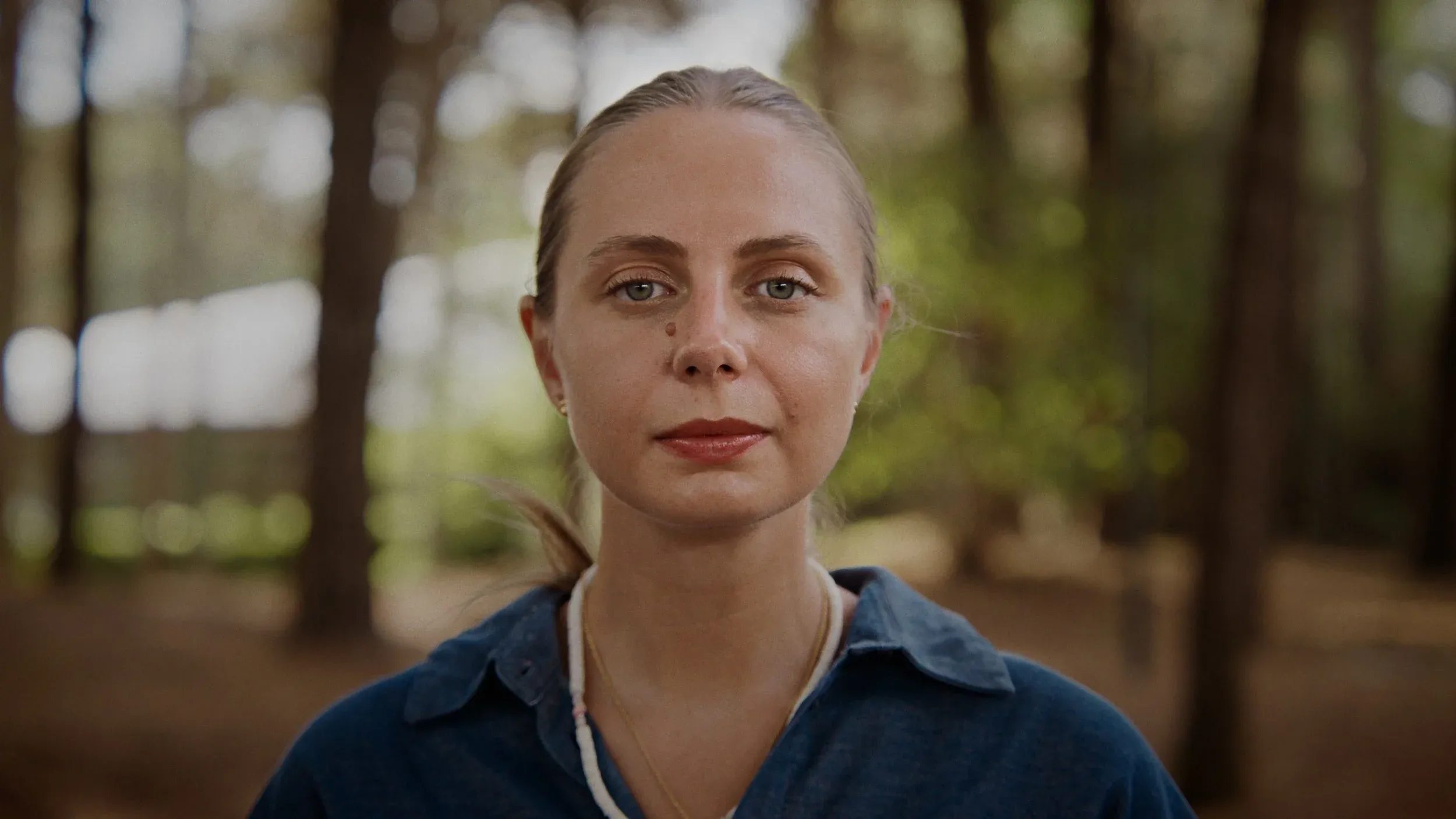 Close-up of the woman in the pine woods looking straight into the camera, both eyes clearly visible
