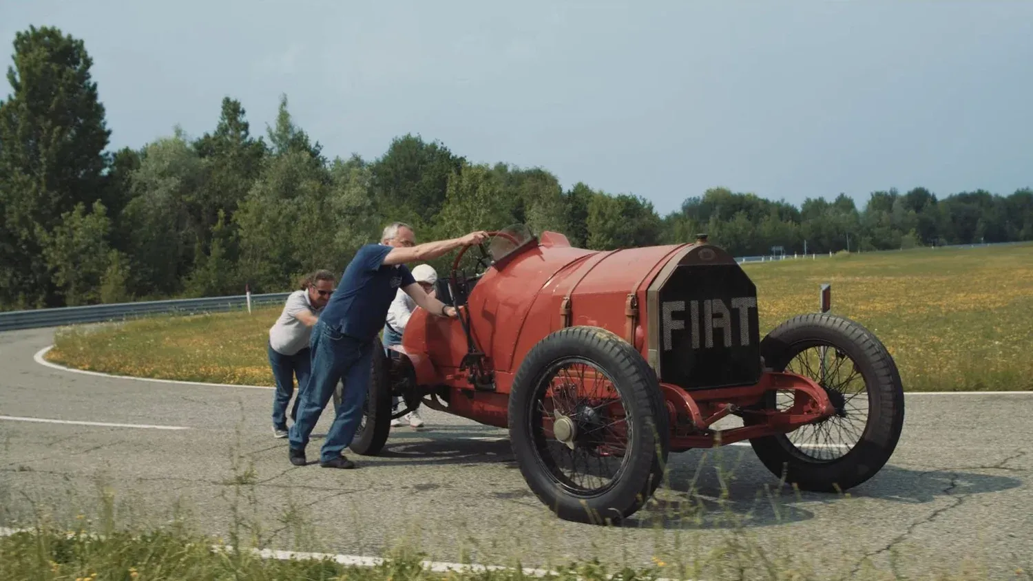 Two men pushing a vintage red Fiat racing car along a track