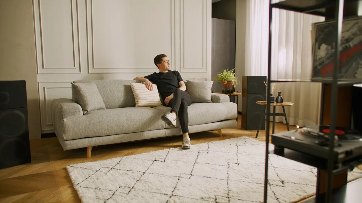 A man sitting on a grey sofa in a living room with a rug and a record player