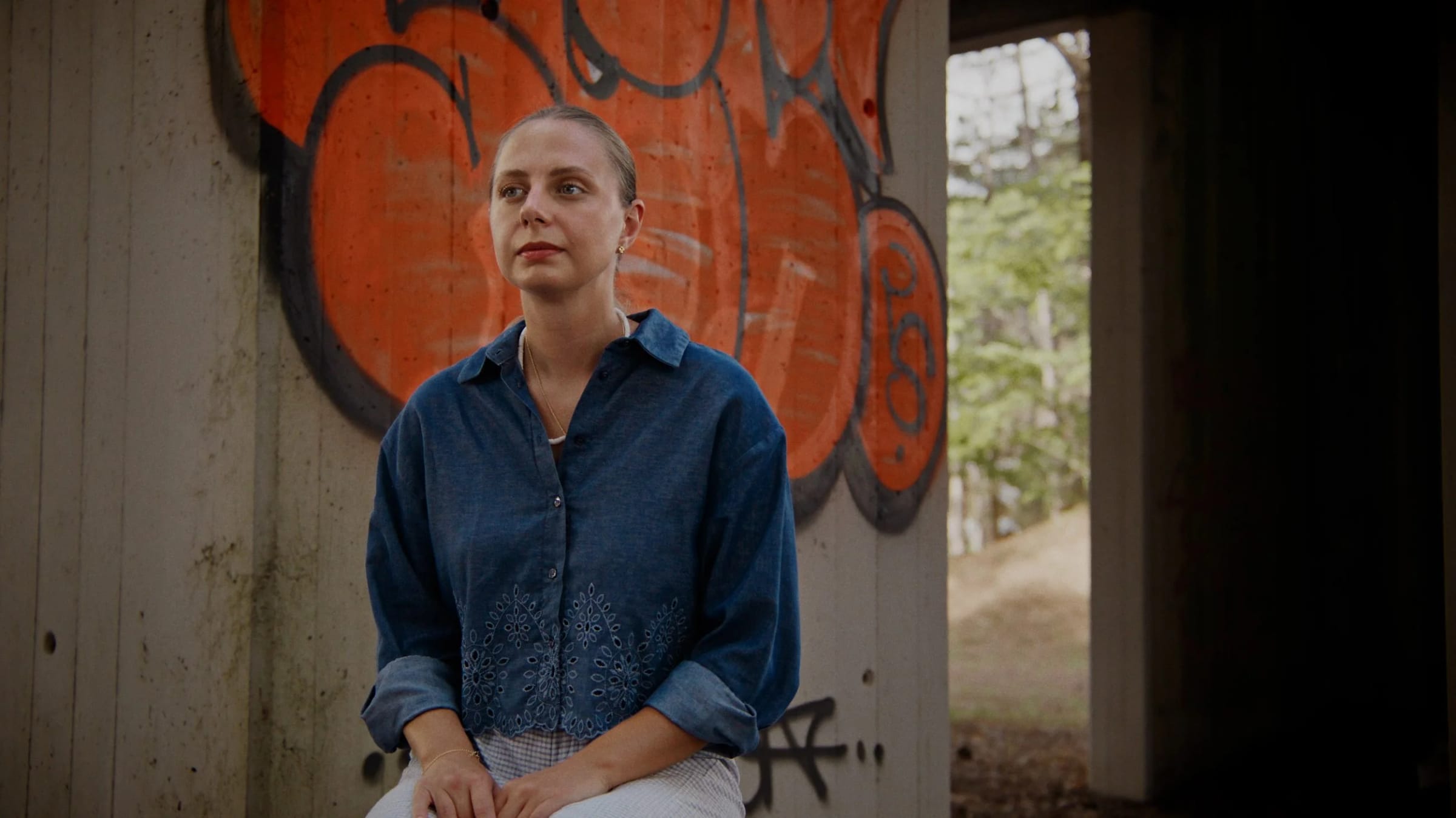 Interview frame: a woman in a denim shirt sits in front of an orange graffiti wall, shot from a low angle