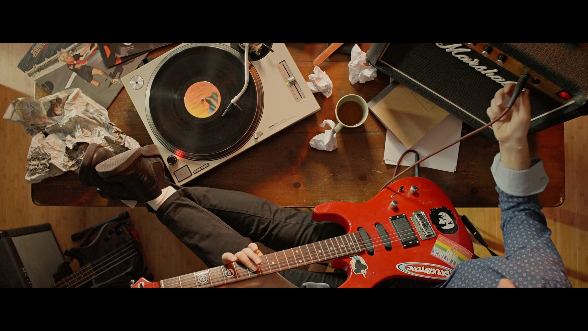 Overhead shot of a cluttered desk with a turntable and a Marshall amp, a hand playing a red electric guitar