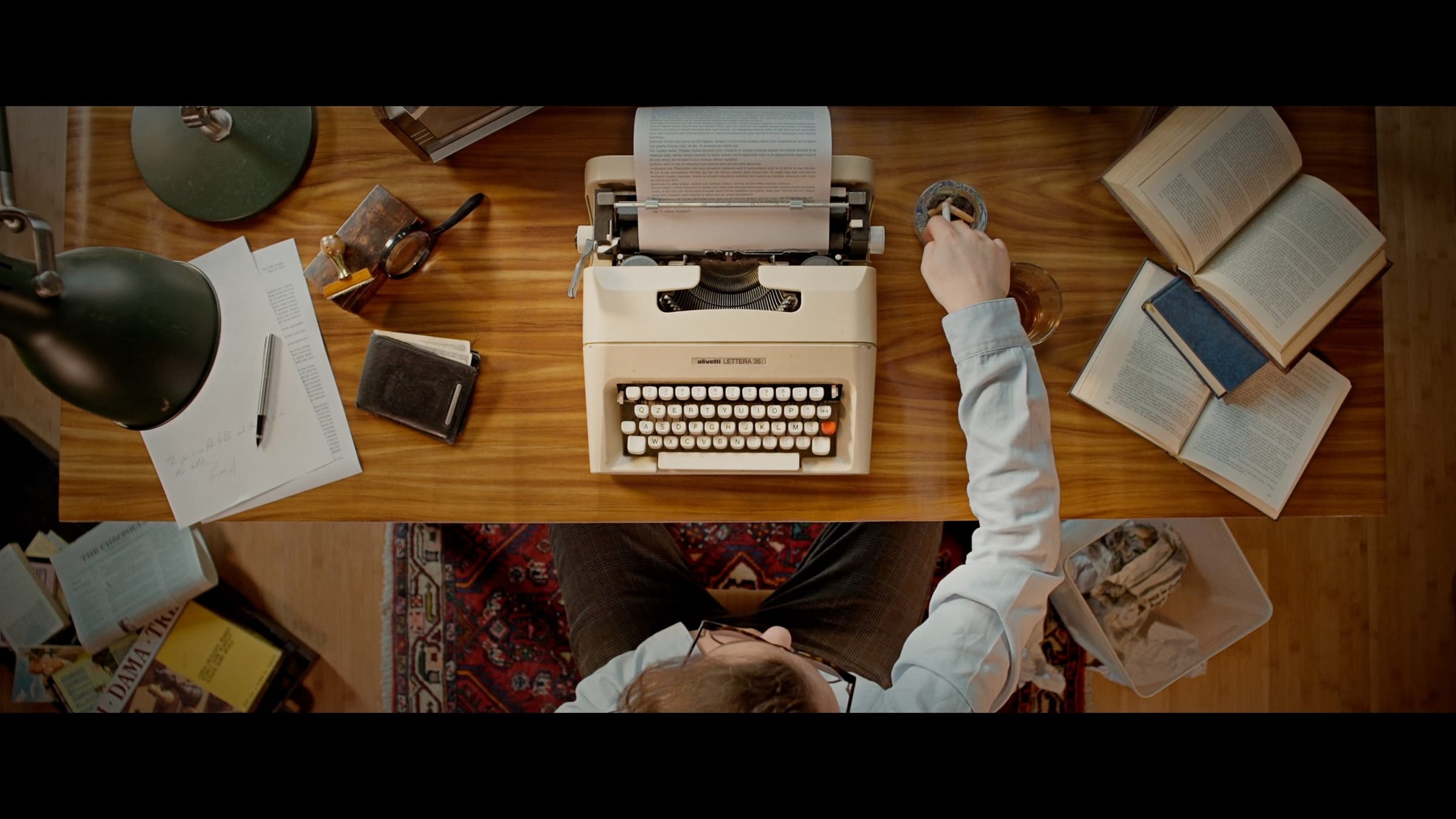 Overhead shot of a man typing on a vintage typewriter at a wooden desk covered with open books