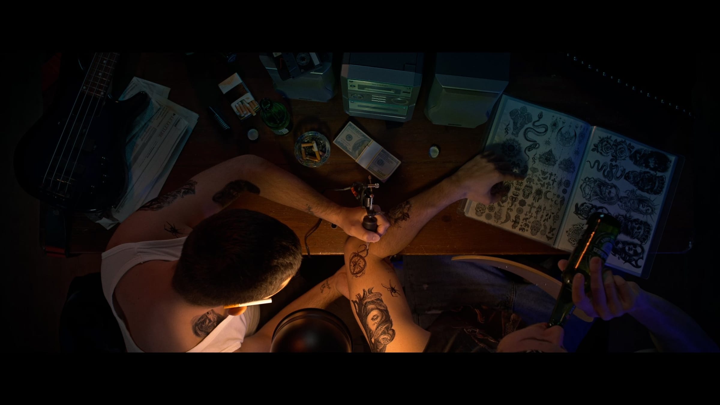 Overhead shot of a tattoo artist working on a client's arm next to a sheet of tattoo designs, lit by a desk lamp